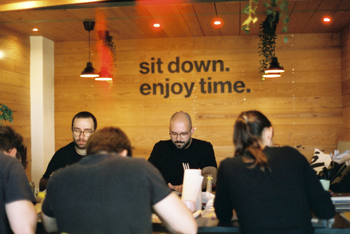 A group of people seated at a wooden table in a restaurant or cafe, with a warm wood-paneled wall behind them featuring the text 'sit down. enjoy time.