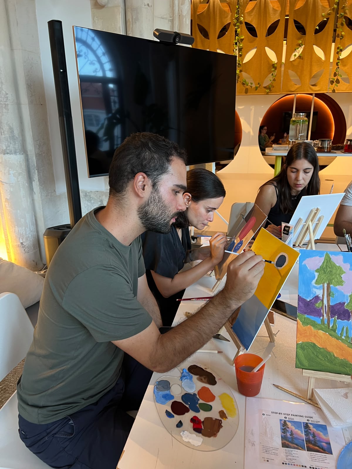 A group of people seated at a table engaging in a painting workshop, with a man in the foreground holding a paintbrush over a canvas and a colorful painted landscape on an easel in front of him.